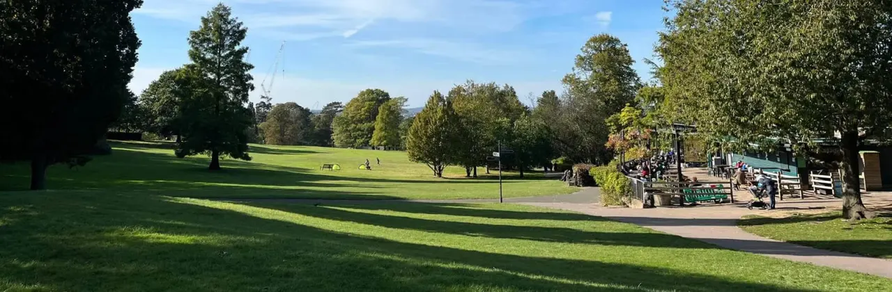 Spacious park with rolling green lawns, mature trees, and visitors gathered near facilities on a sunny day.