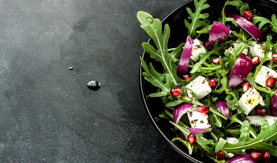 This is a PHOTOGRAPH.

Salad in a black bowl with rocket, purple onion, white cheese and pomegranate seeds on a dark surface.