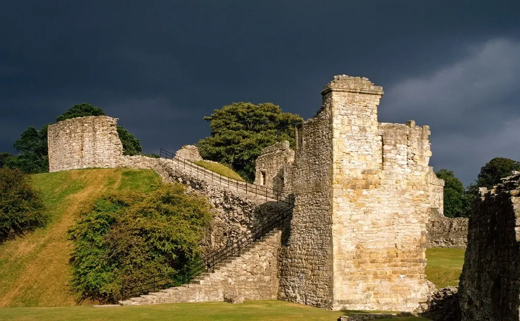 Medieval castle ruins with stone towers and fortified walls surrounded by grassed grounds under dramatic sky.
