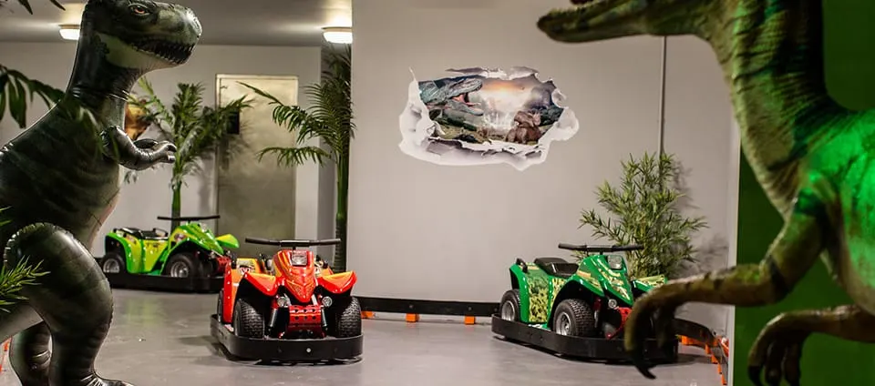 Dinosaur models flanking a ride-on vehicle experience with red and green electric cars on a track indoors.