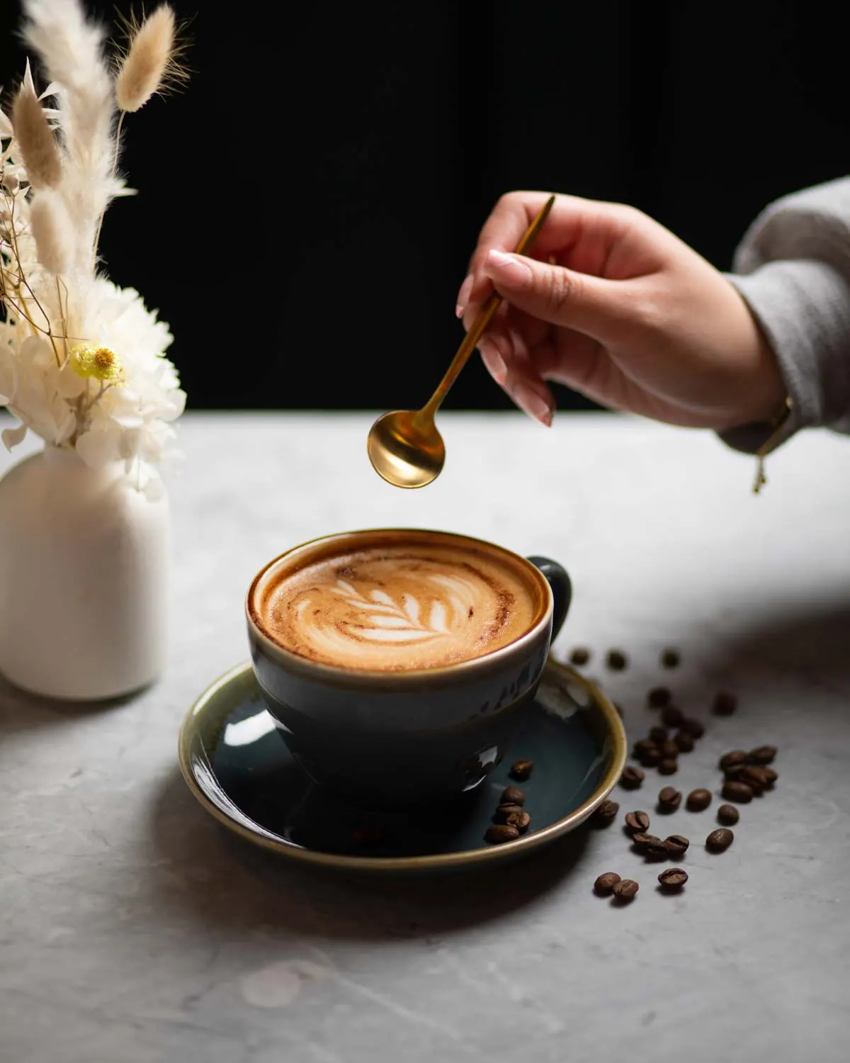 Person drizzling honey into a cappuccino with latte art, surrounded by coffee beans and white flowers.