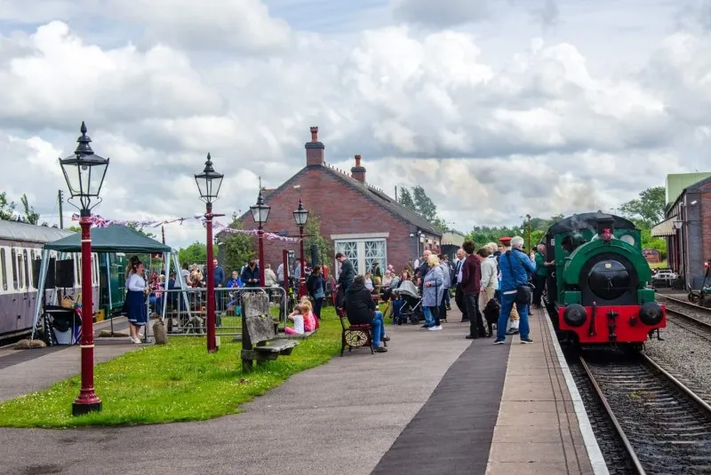 Visitors stroll around a heritage railway station with a red steam locomotive, Victorian buildings, and vintage street lamp on a cloudy day.