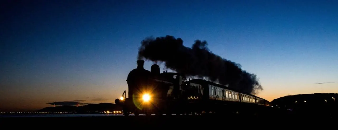 Steam locomotive with illuminated headlight travelling at dusk, silhouetted against twilight sky with black smoke billowing from chimney.
