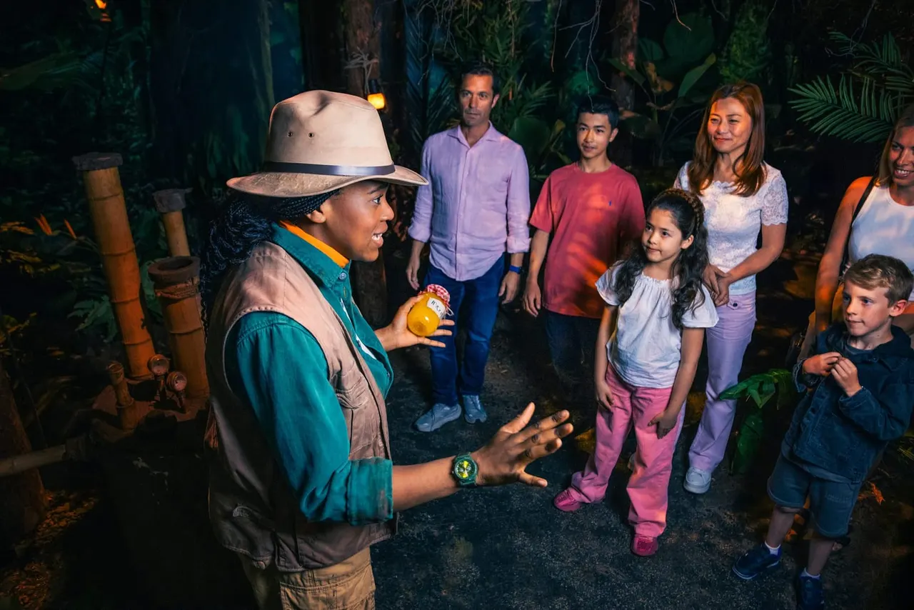A uniformed guide speaks to a group of adults and children in a tropical forest setting at night.