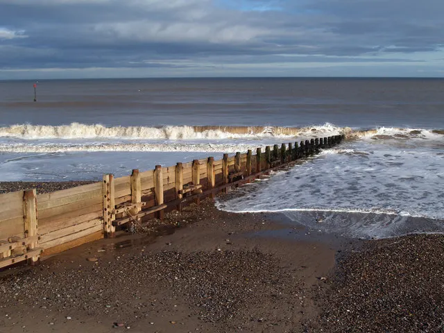 Wooden groynes extending into the sea with breaking waves on a pebbly beach under a cloudy sky.