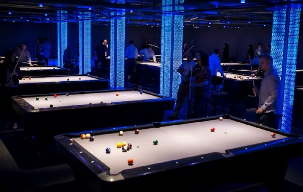 Pool tables with coloured balls set up in a modern venue with blue lighting and several people playing or socialising.