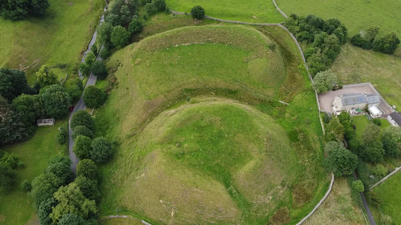 Aerial view of hillfort earthworks with concentric circular ramparts, surrounded by grassland, trees, and a modern building.