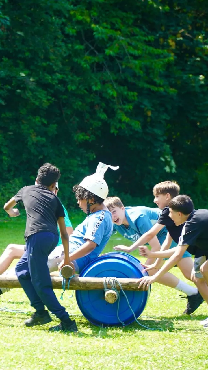 A group of children work together to push a large blue barrel across grass during an outdoor team activity.