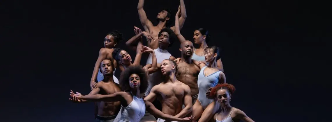 Group of dancers performing an ensemble contemporary dance piece on stage against a black background.
