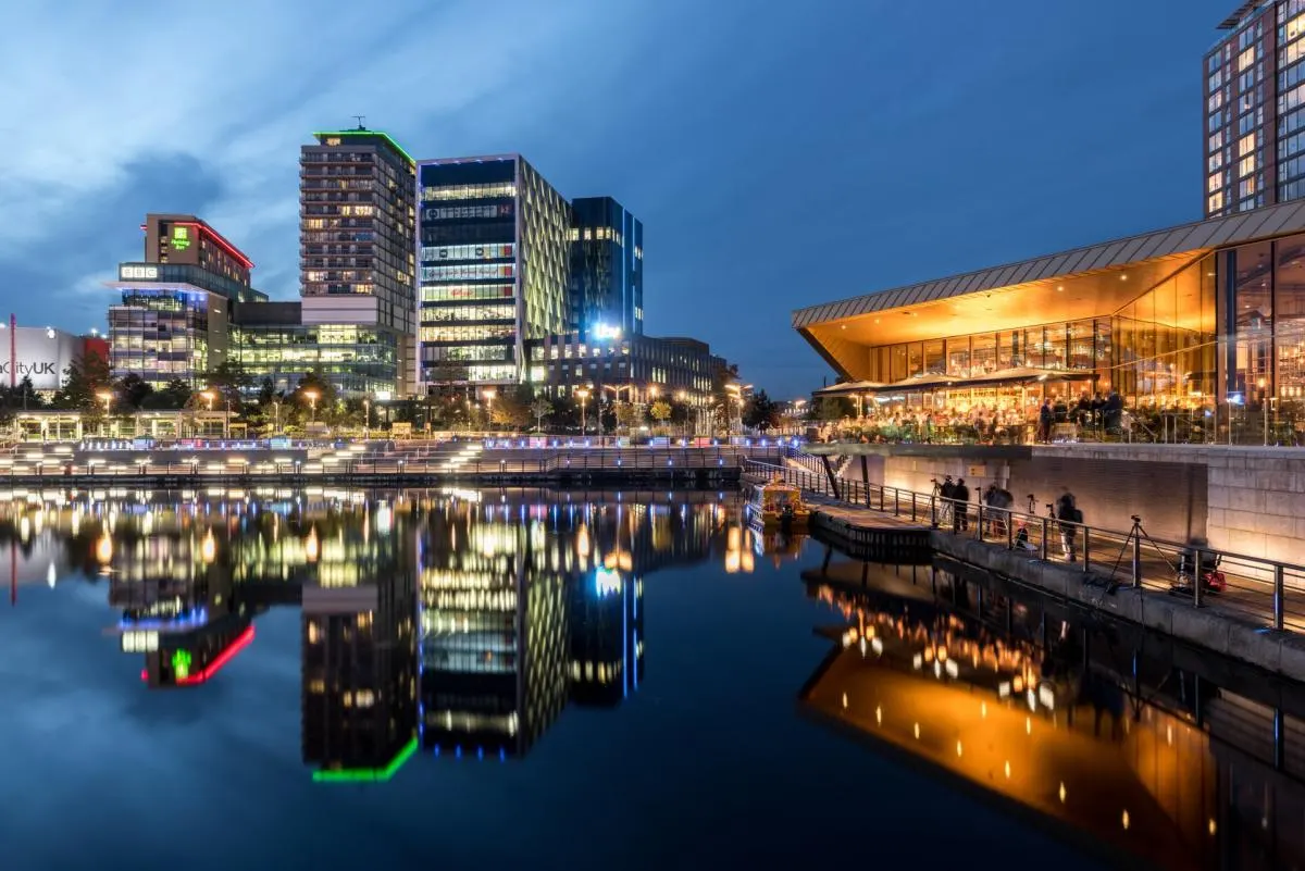 Illuminated waterfront at dusk with modern buildings reflected in still water, people on a promenade, and a contemporary curved structure on the right.