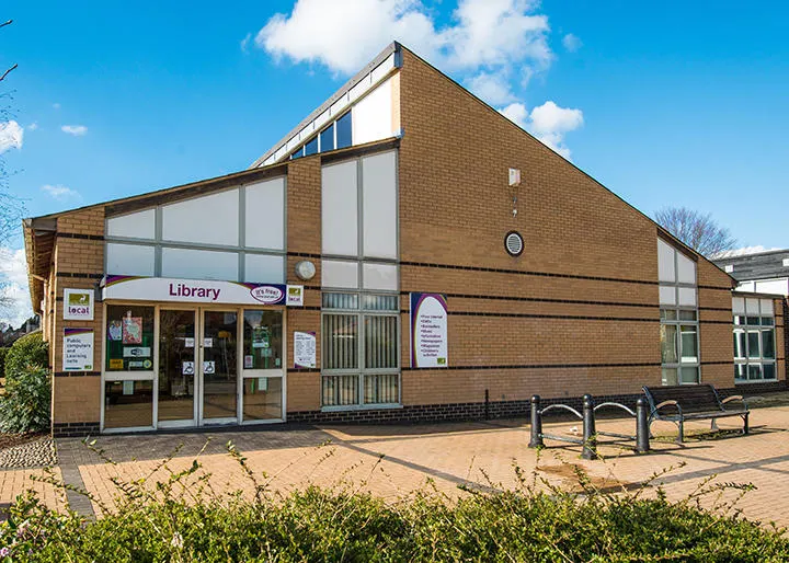 Modern brick library building with large windows and entrance doors, set in a paved courtyard with benches under blue sky.