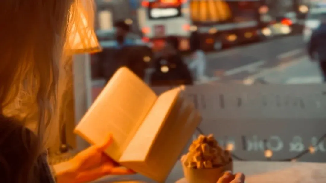 Person reading a book by a window with a coffee cup, urban street scene visible outside.