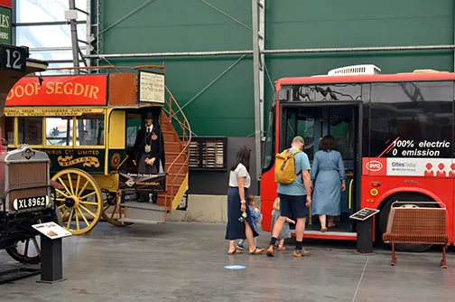 Vintage yellow horse-drawn cart and red electric bus with visitors exploring them at an indoor museum venue.