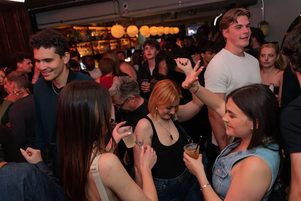 A crowd of young adults dancing and socialising in a dimly lit bar venue with warm ambient lighting.