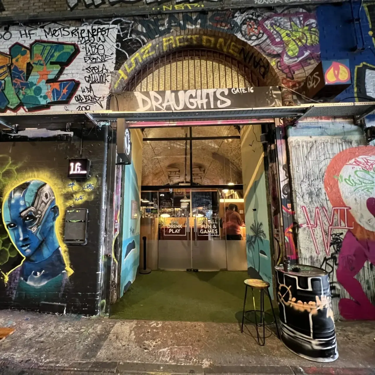 Street-level entrance to a games bar with colourful graffiti art on surrounding walls and open glass doors.