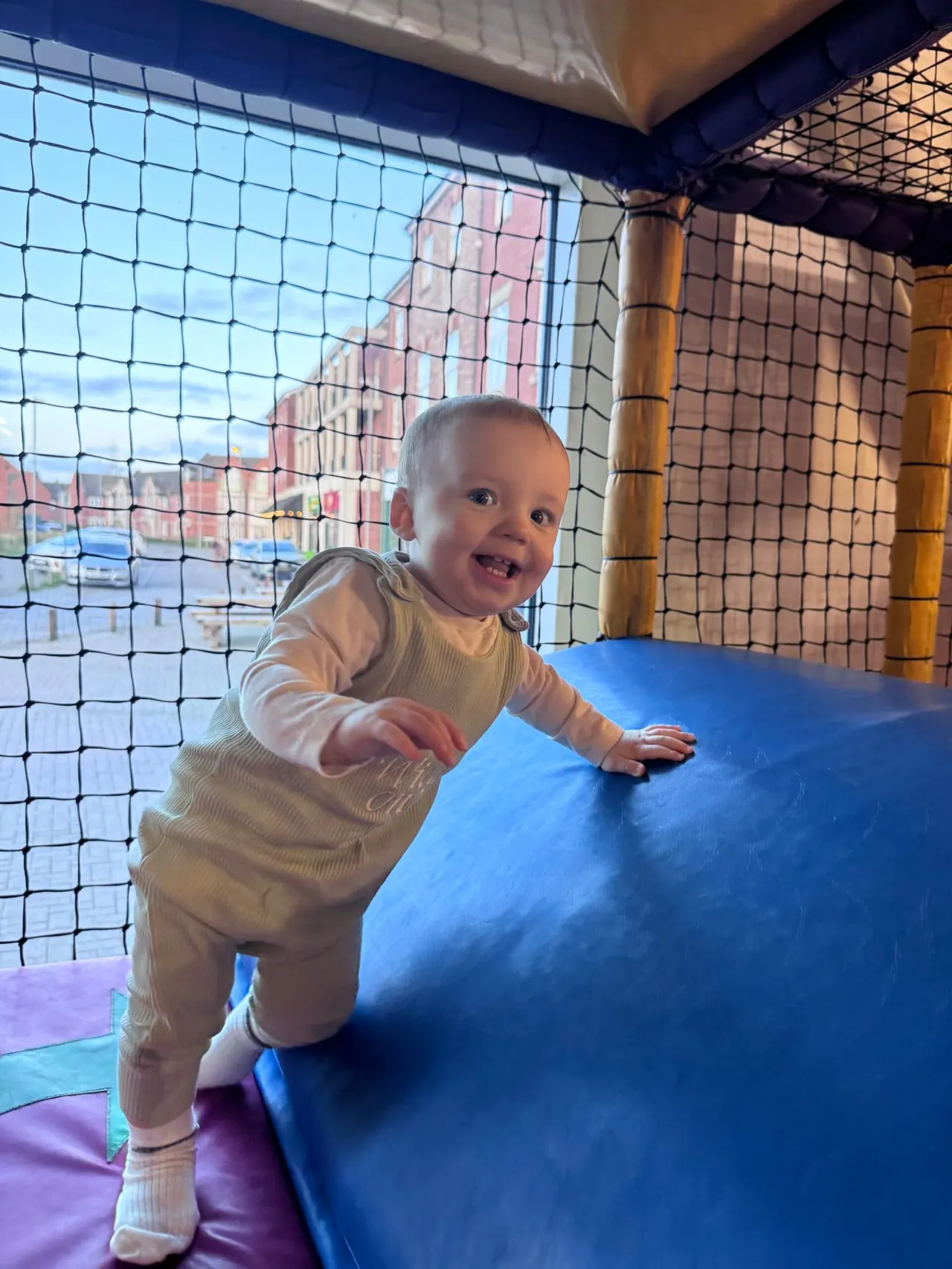 Toddler playing on a blue foam structure in an indoor soft play area with safety netting.