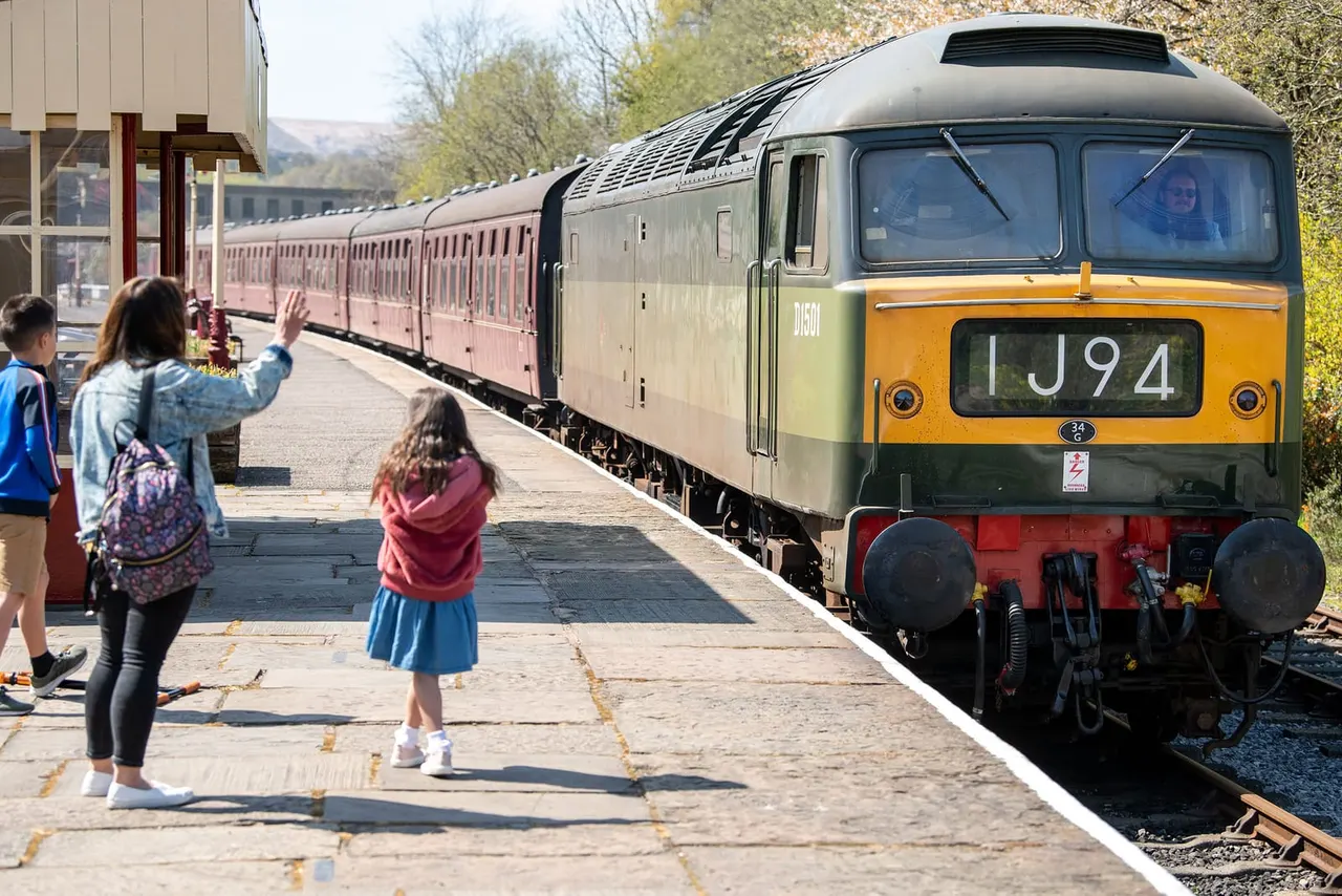 Children on a railway platform waving at a vintage diesel locomotive and passenger train at a heritage railway station.