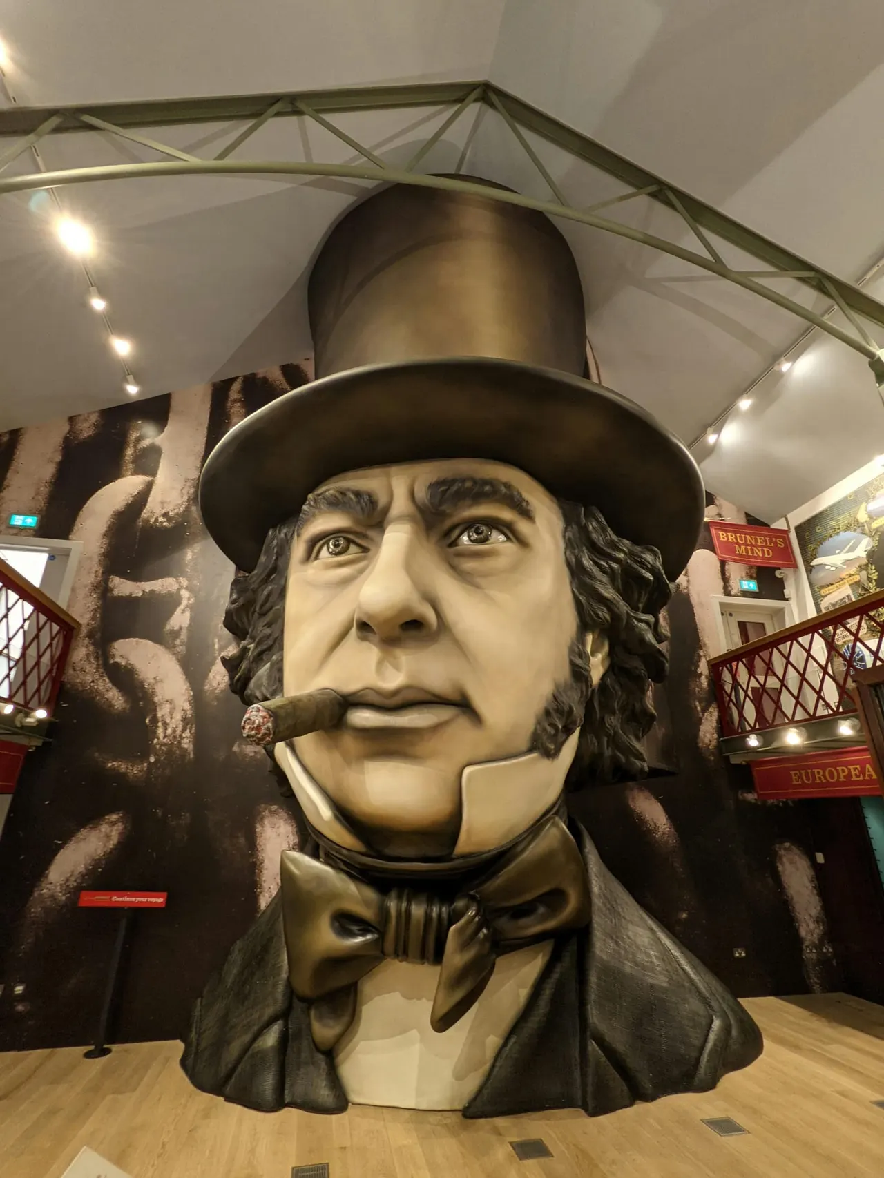 Large sculptural bust of a man wearing a top hat and smoking a cigar, displayed indoors under modern lighting and architectural framework.