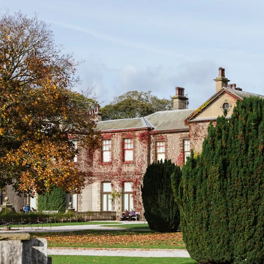 Two-storey brick country house with ivy-covered walls, surrounded by formal gardens with topiary hedges in autumn.