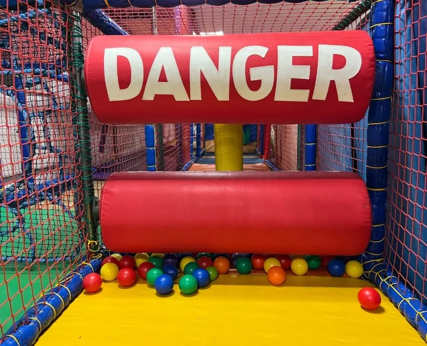 Inflatable obstacle course with red "DANGER" sign and coloured balls inside a mesh-enclosed play structure.
