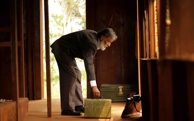 Man in dark suit bending over a wooden box in a gallery doorway with natural light behind.