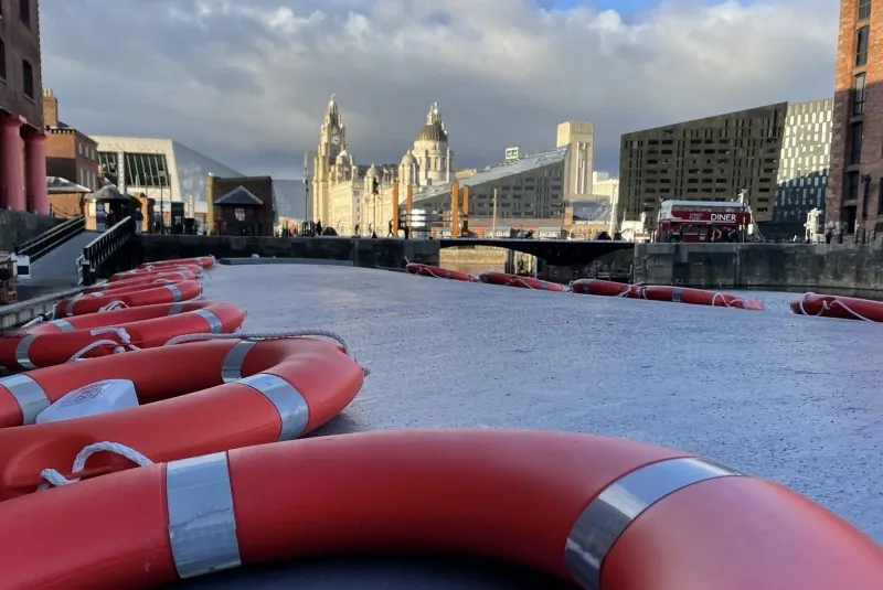 Red inflatable buoys arranged on a boat deck with Liverpool's waterfront and historic buildings visible across the water.