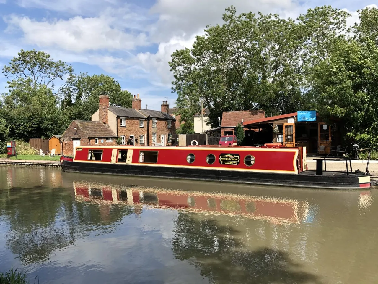 Red and cream narrowboat moored on a canal beside brick buildings and trees.