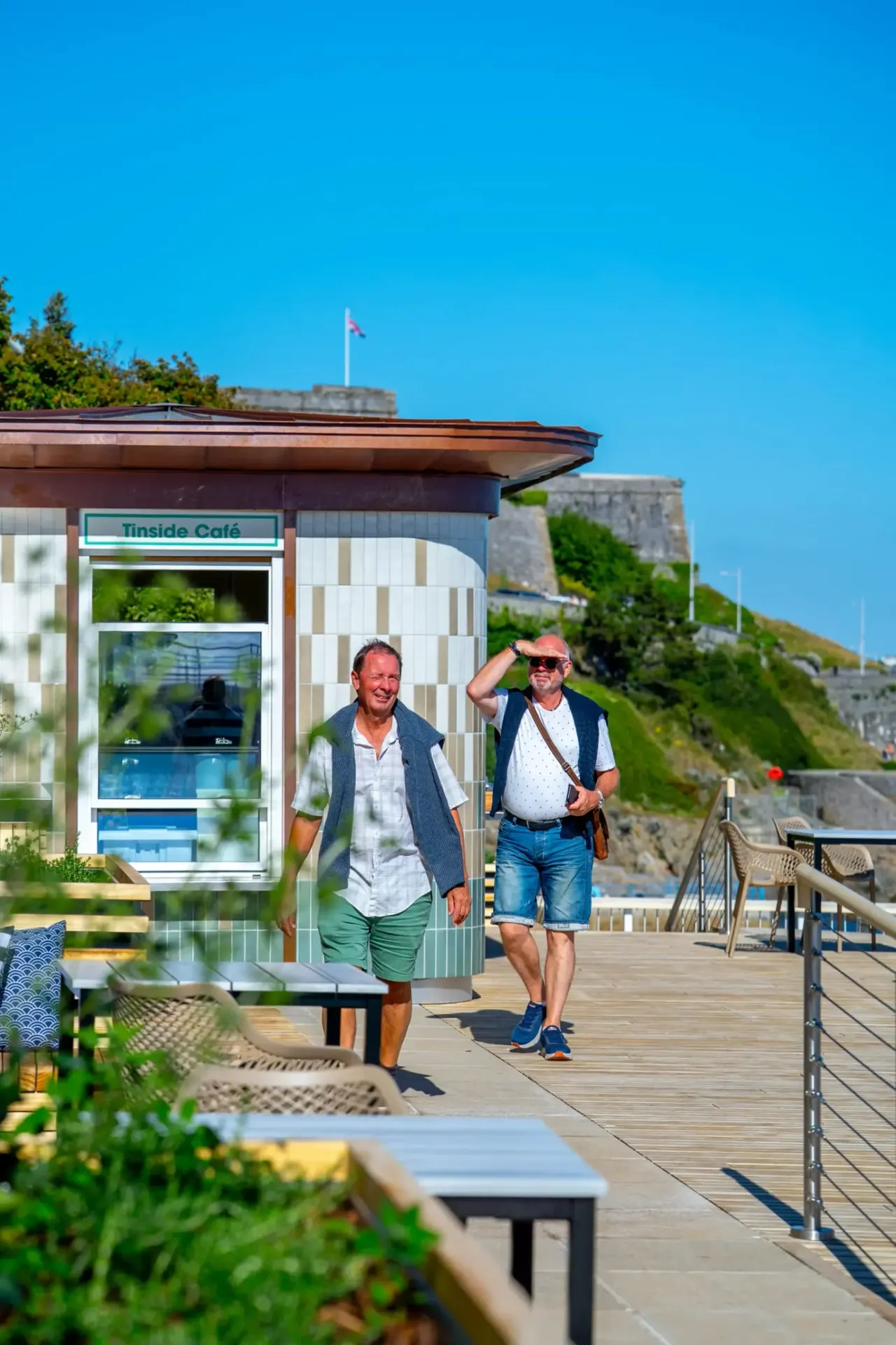 Two men walk past a seaside café kiosk on a wooden promenade under clear blue sky, with fortified walls visible behind them.