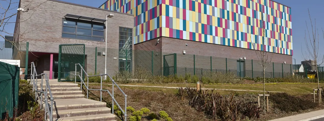 Modern brick leisure building with colourful striped panels, metal stairs with railings leading to entrance, and landscaped grounds with shrubs.