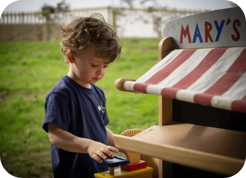 Young child in navy shirt playing at a wooden toy shop counter with striped awning in a garden setting.