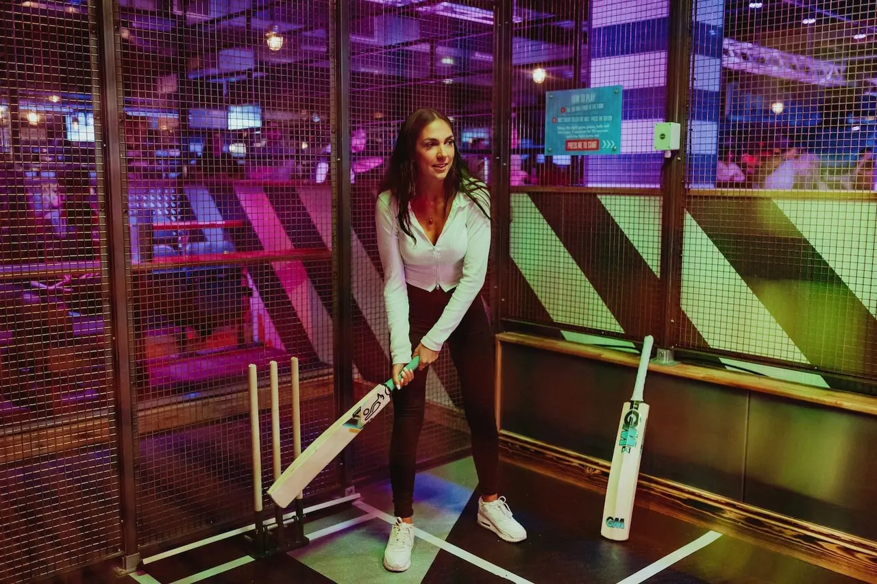 Woman holding a cricket bat at an indoor cricket net with purple and green neon lighting.