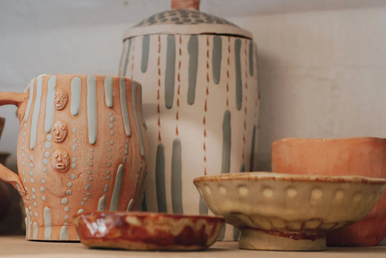 Handmade ceramic vessels in earth tones displayed together, including an orange jug with face motifs and a lidded jar with striped patterns.