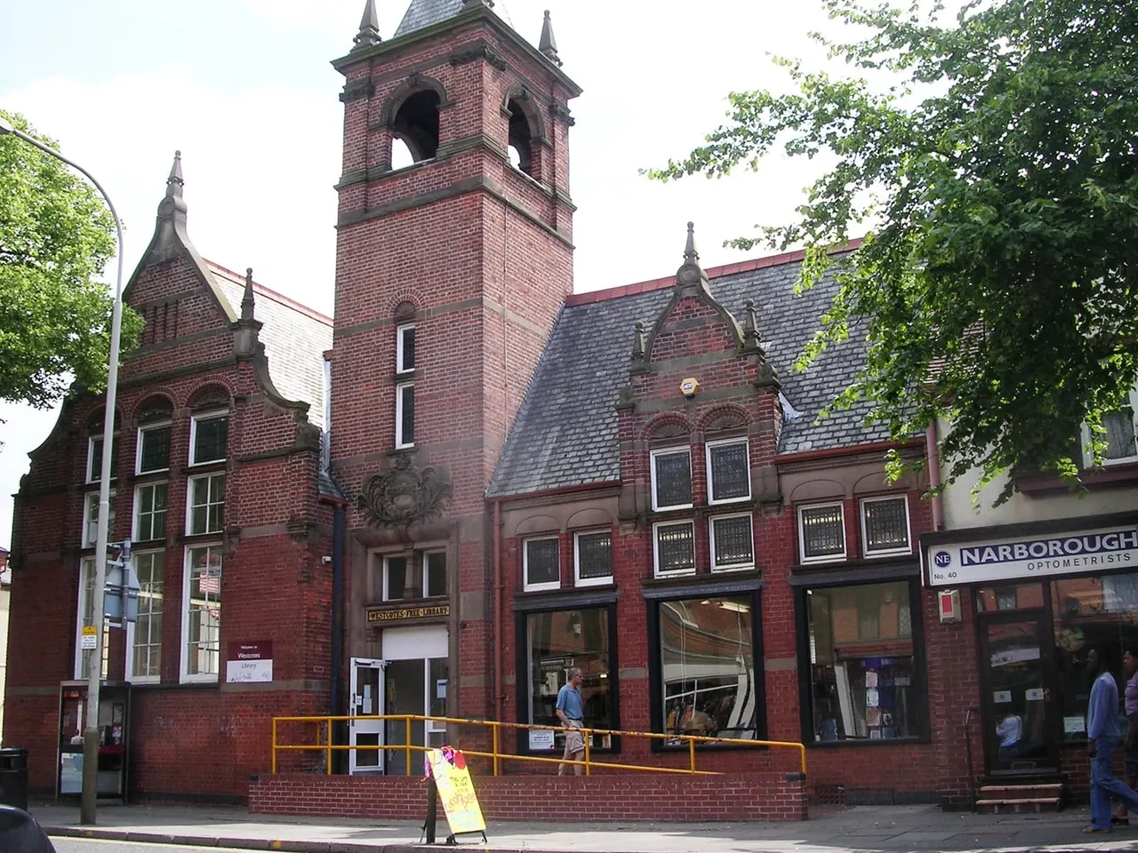 Victorian red-brick library building with a central bell tower, street-level shopfronts, and people on the pavement outside.