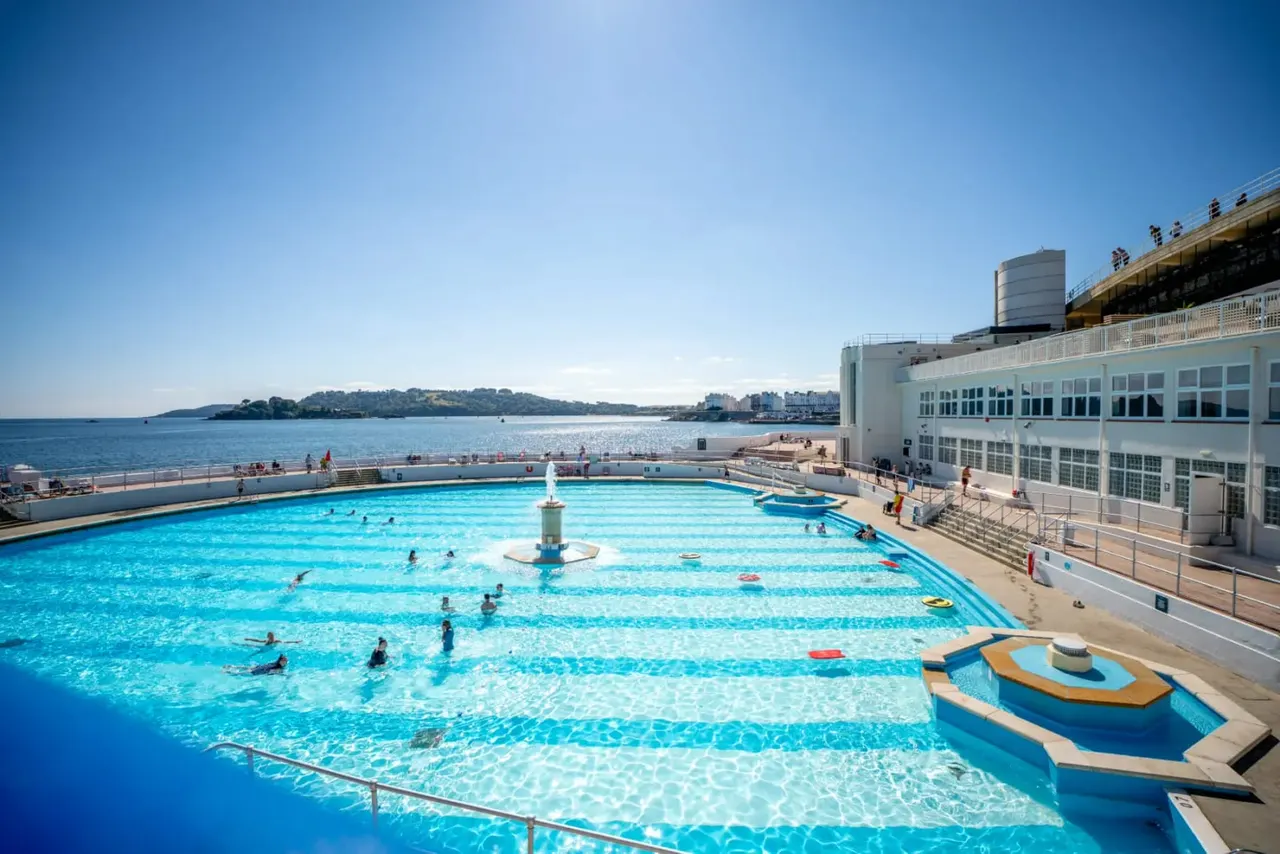 Large outdoor swimming pool with bathers, overlooking a bay with islands and coastal buildings under clear blue sky.