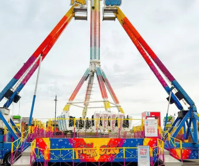 Colourful fairground swing ride with rainbow-striped support arms and bright red and yellow decorated cabin, set against a cloudy sky.