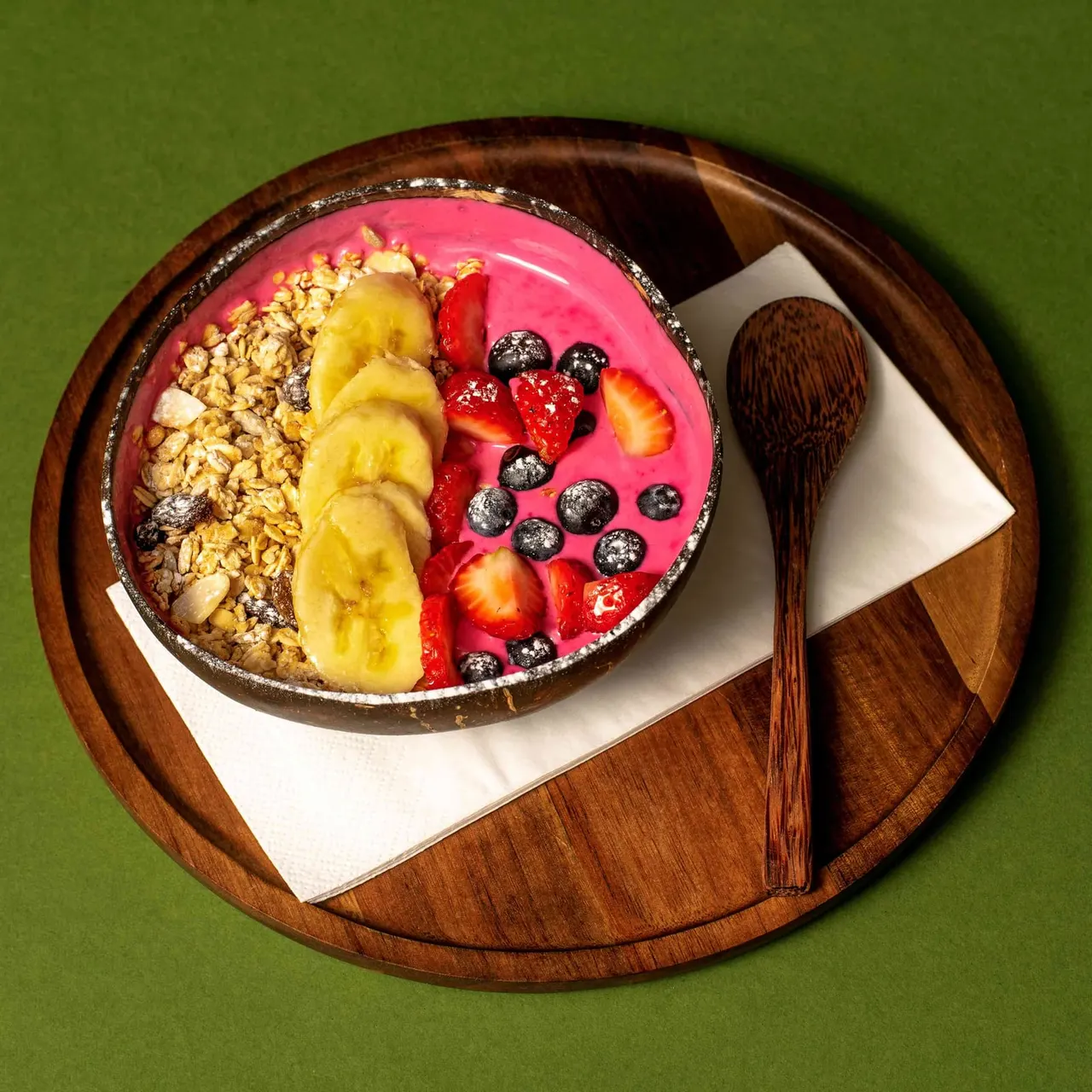 Açai bowl with granola, banana slices, berries and coconut served on wooden board with spoon.