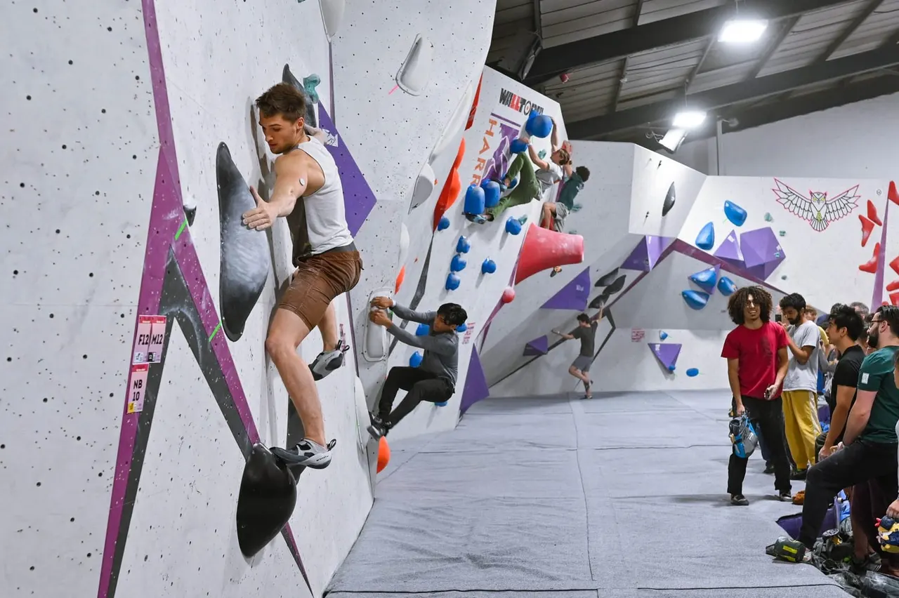 Young man climbing an indoor wall with coloured holds whilst others watch from the ground floor.