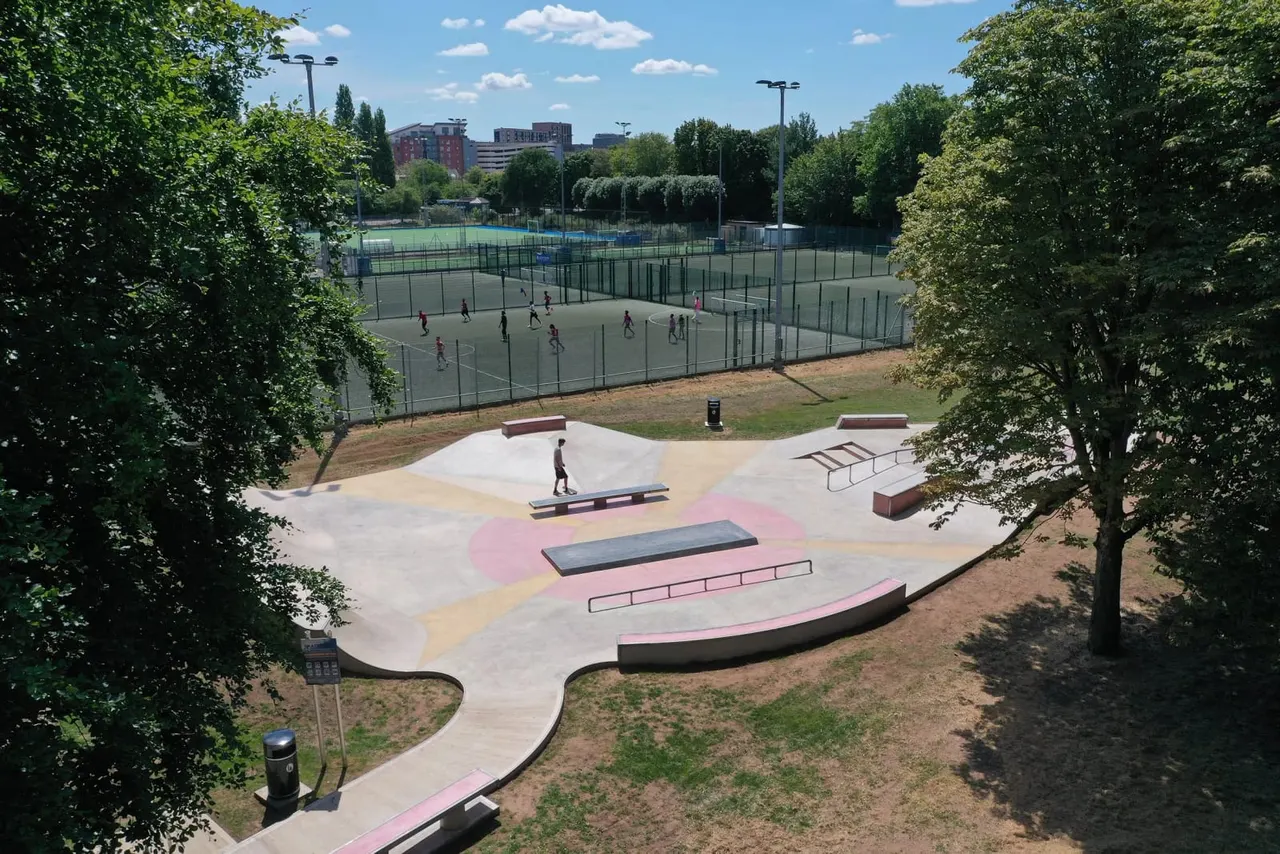 A young skateboarder practising on a modern skate park with ramps and rails, surrounded by trees and sports courts in the background.