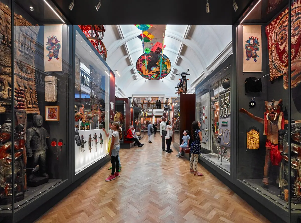 Visitors of various ages explore a museum gallery displaying Asian artefacts, sculptures and textiles in illuminated display cases beneath a skylighted ceiling.