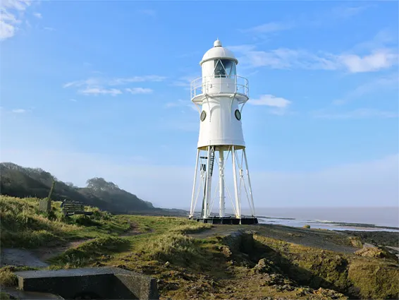 White lighthouse standing on rocky shore with green hillside and water in background under blue sky.