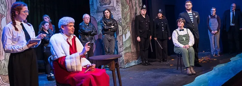A theatrical cast performs a scene on stage with multiple actors in various costumes under blue lighting.