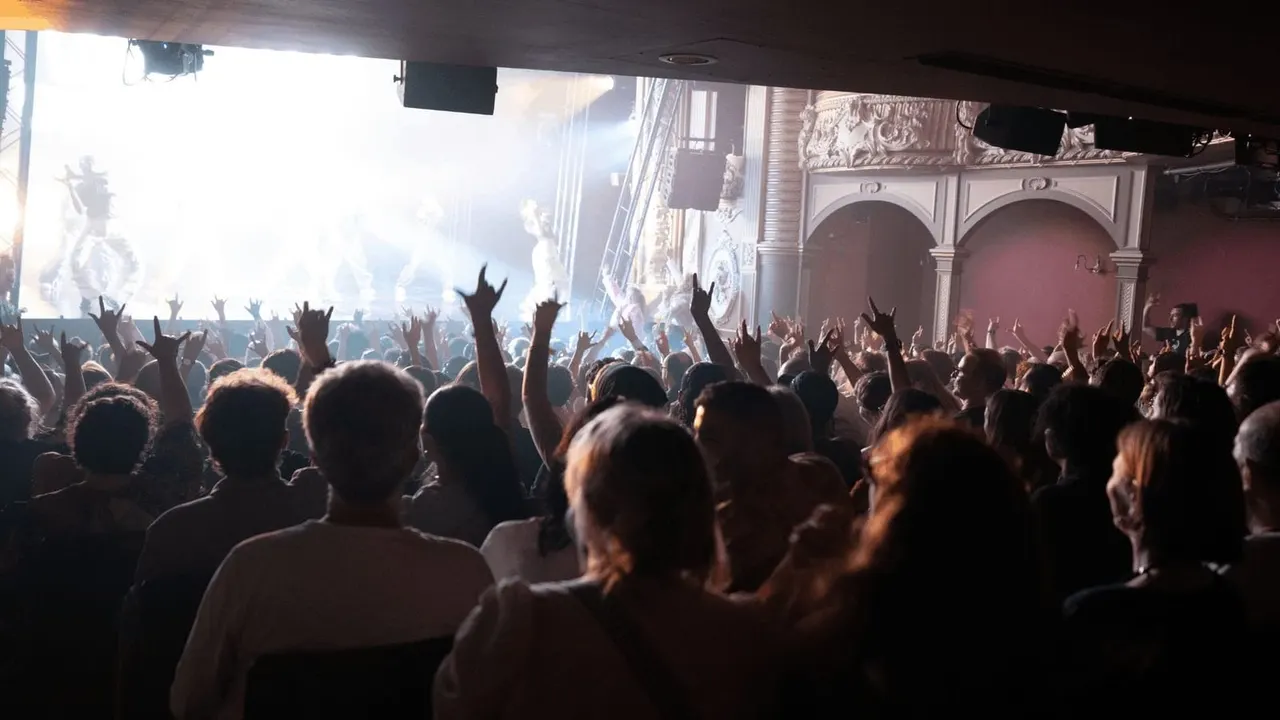 Large crowd with raised arms watching a concert or live performance on stage with bright lighting.