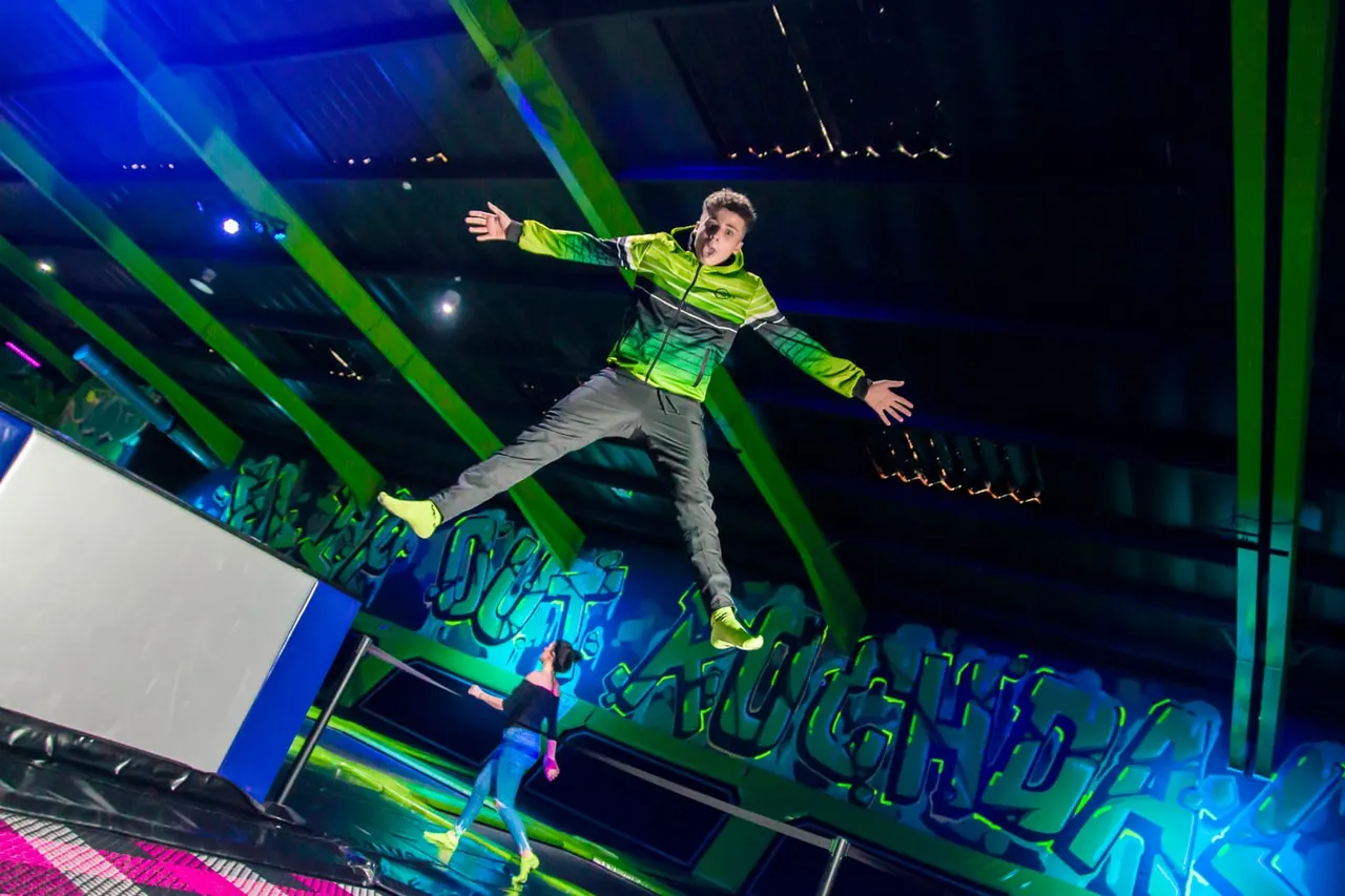 Person in neon yellow jacket leaping between green padded obstacles in an indoor trampoline park with blue lighting.
