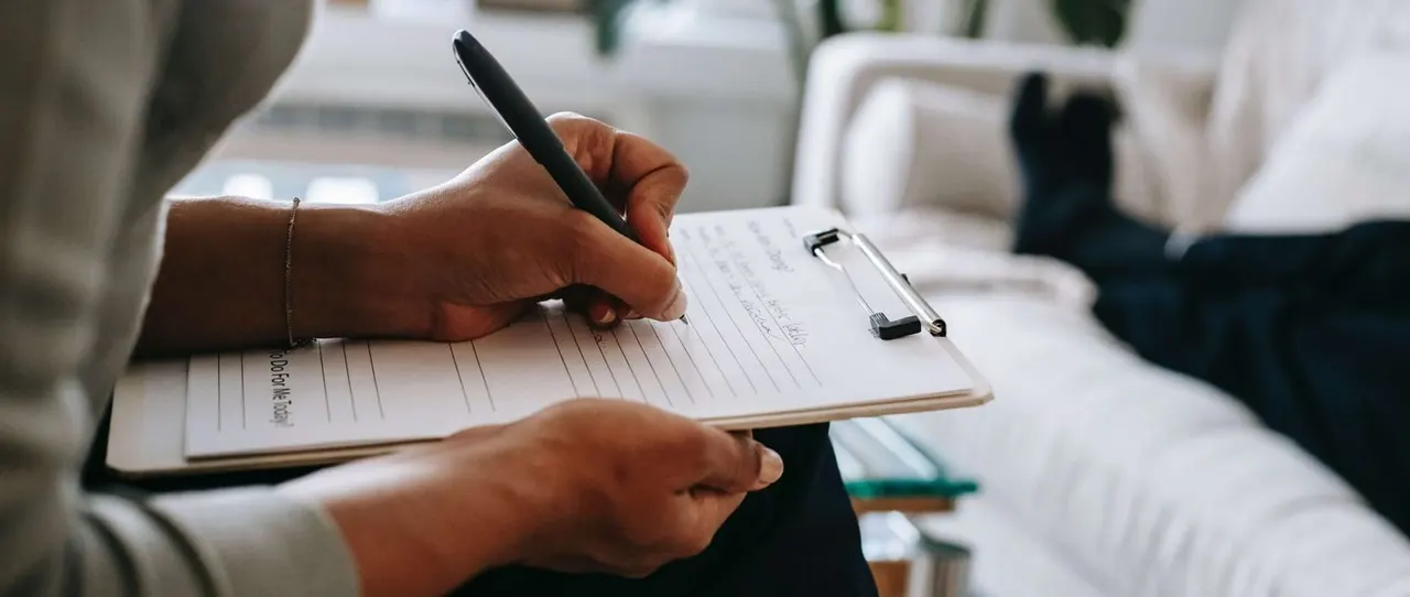 Person writing on a clipboard with a pen in an indoor setting with potted plants.