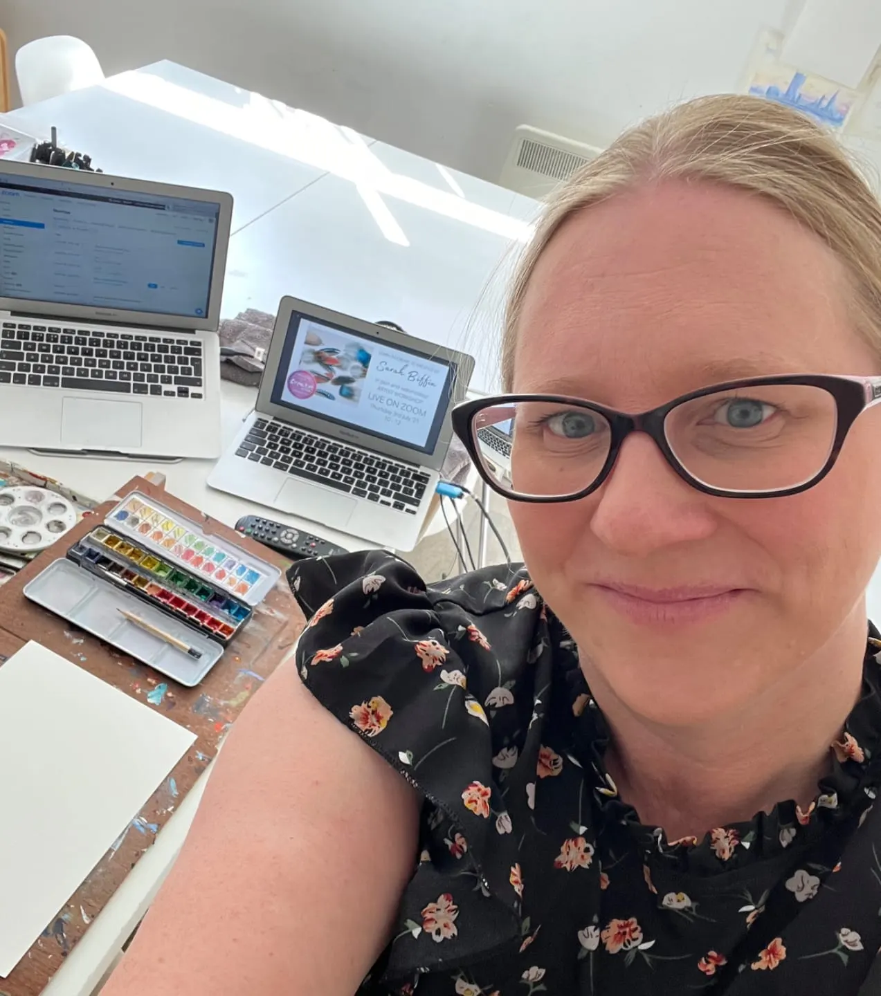 Woman wearing glasses and floral top takes selfie at desk surrounded by laptops and watercolour paints.
