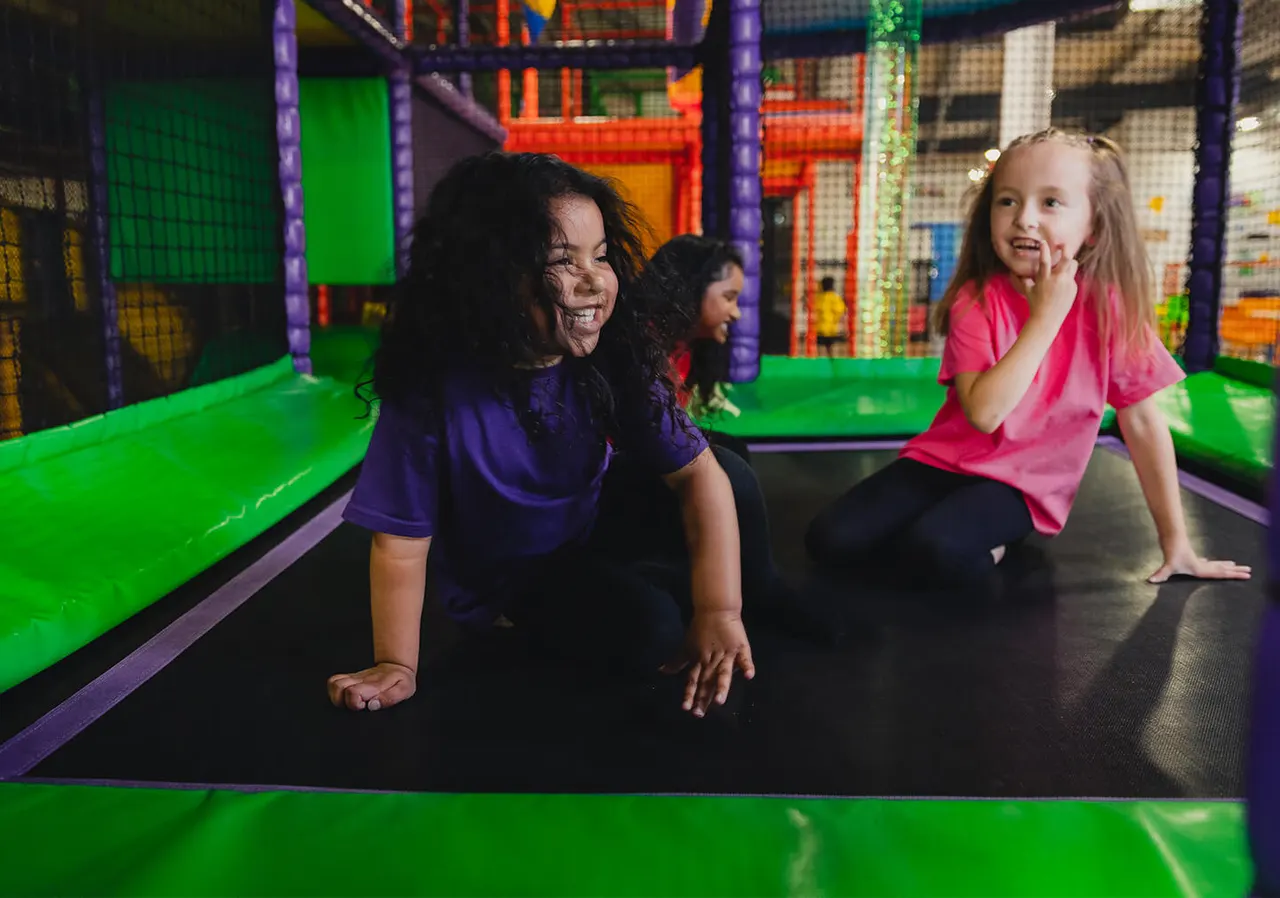 Children playing and jumping on a brightly lit indoor trampoline with coloured netting and foam structures.