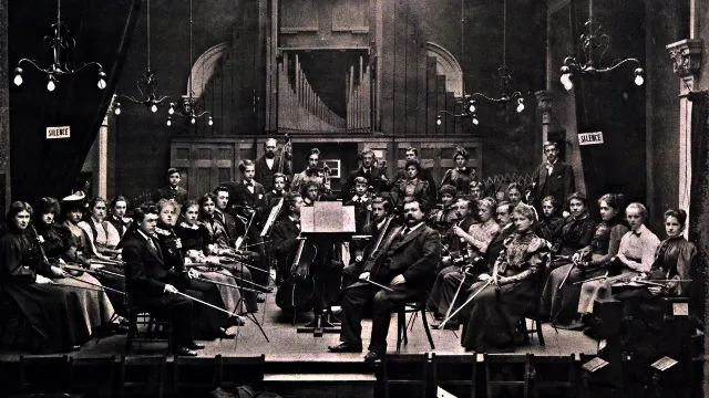 Large orchestra and conductor performing in a grand concert hall with ornate architecture and hanging chandeliers.
