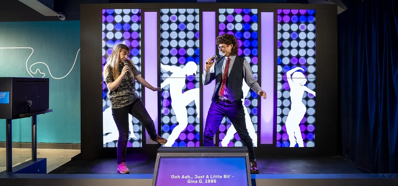 Two visitors perform a duet with microphones on stage in front of an illuminated backdrop displaying silhouettes of dancers.