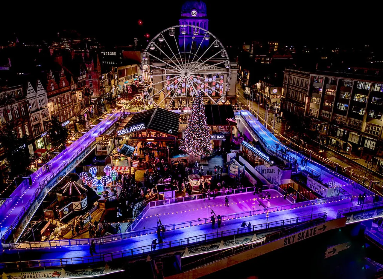 Night-time aerial view of an illuminated winter fair with a large Ferris wheel, ice rink, festive market stalls, and crowds enjoying seasonal attractions.
