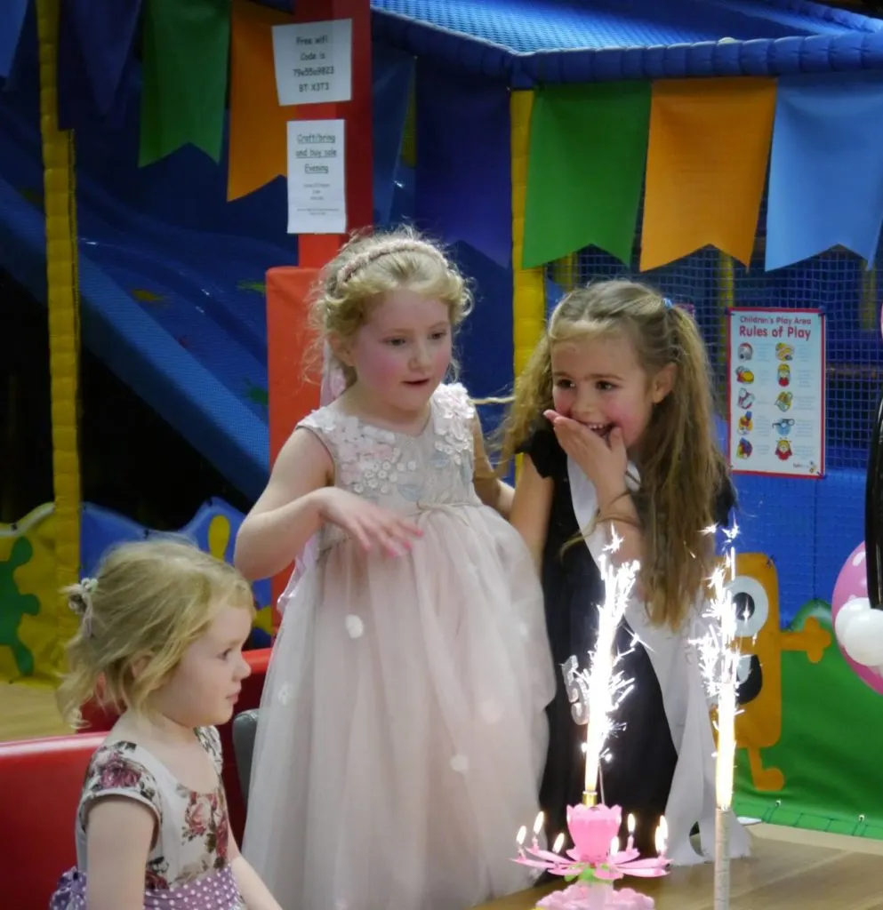 Three young girls gathered around a birthday cake with sparkler candles in a colourful soft play arena.